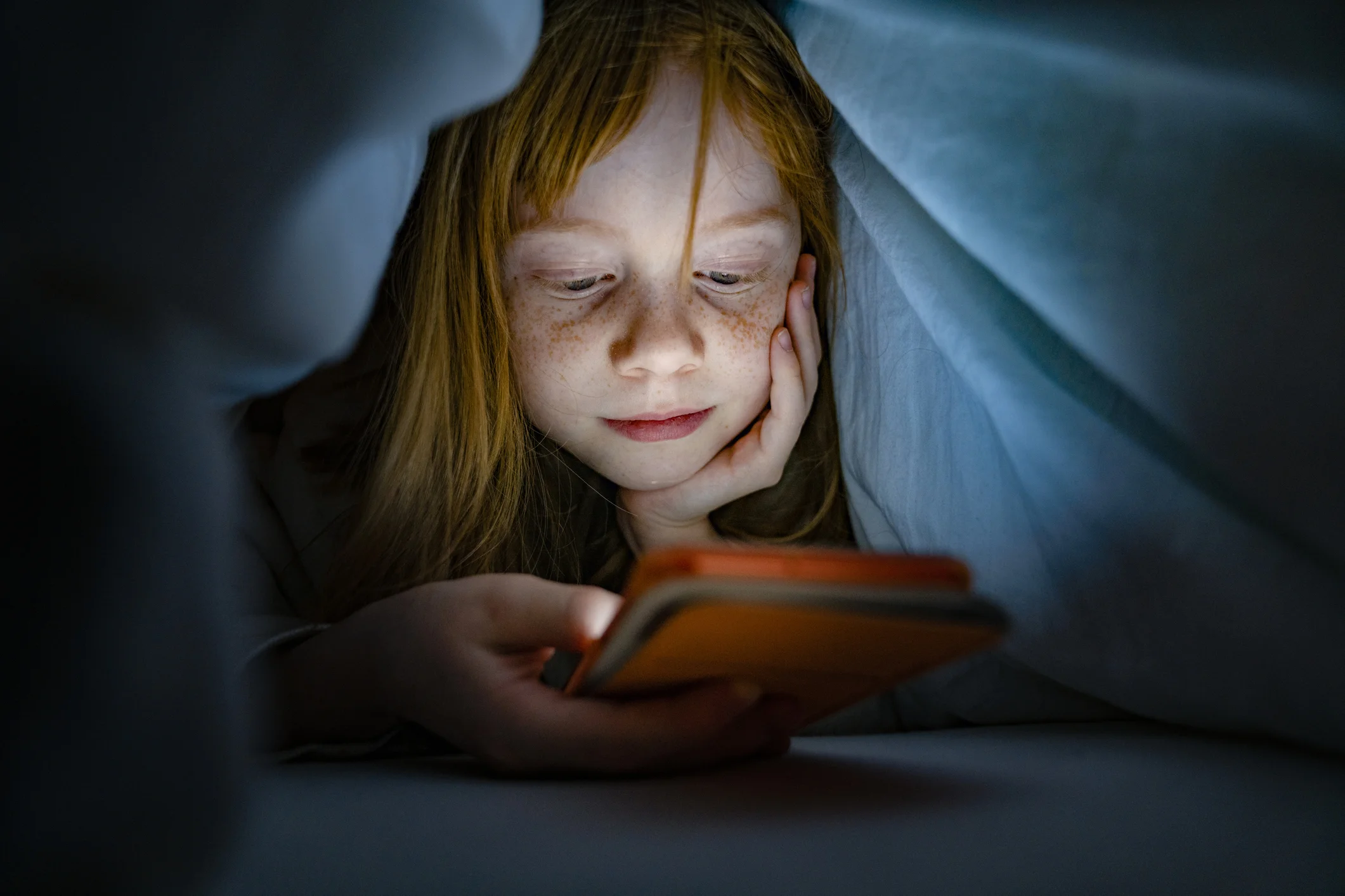 Una bambina guarda uno smartphone sotto le coperte